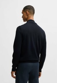 Maglione nero a maglia con colletto alto, maniche lunghe e polsini aderenti. Tessuto liscio con una leggera lucentezza, indossato sopra pantaloni scuri.