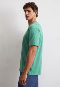 Zielona bawełniana koszulka z krótkim rękawem, okrągłym dekoltem i emblematem w pobliżu ramienia; zestawiona z niebieskimi dżinsowymi szortami.