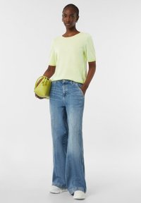 Lichtgroene, gestreepte gebreide trui gecombineerd met blauwe wide-leg jeans. Houdt een kleine, ronde, groene handtas vast. Witte sneakers maken de outfit af.