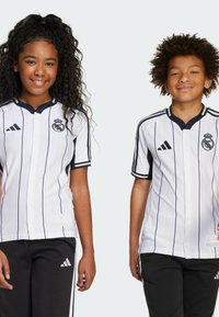 Magliette bianche a maniche corte con strisce navy, dotate di chiusura con bottoni, rifiniture nere e logo del Real Madrid. Pantaloni neri con dettagli bianchi.