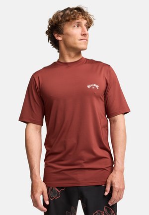Man met krullend haar die een roestkleurig Billabong T-shirt en zwarte shorts met rode bloemmotieven draagt, kijkend naar zijn rechterzijde.