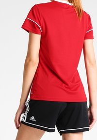 Röd sporttröja med vita detaljer, korta ärmar, rund hals, tillsammans med svarta shorts med vita ränder och logodetaljer.