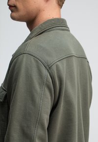 Olijfgroene jas van een zachte, gestructureerde stof. Heeft een kraagontwerp en zichtbare stiksels langs de naden. Zijaanzicht van het kledingstuk.