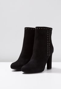Botines de ante negro con tacón alto, que presentan una fila de tachuelas metálicas a lo largo del lado superior para un detalle adicional.