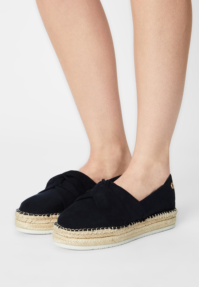 Pieds portant des espadrilles noires slip-on avec un détail noué et des semelles plateforme en jute tressé sur fond blanc.
