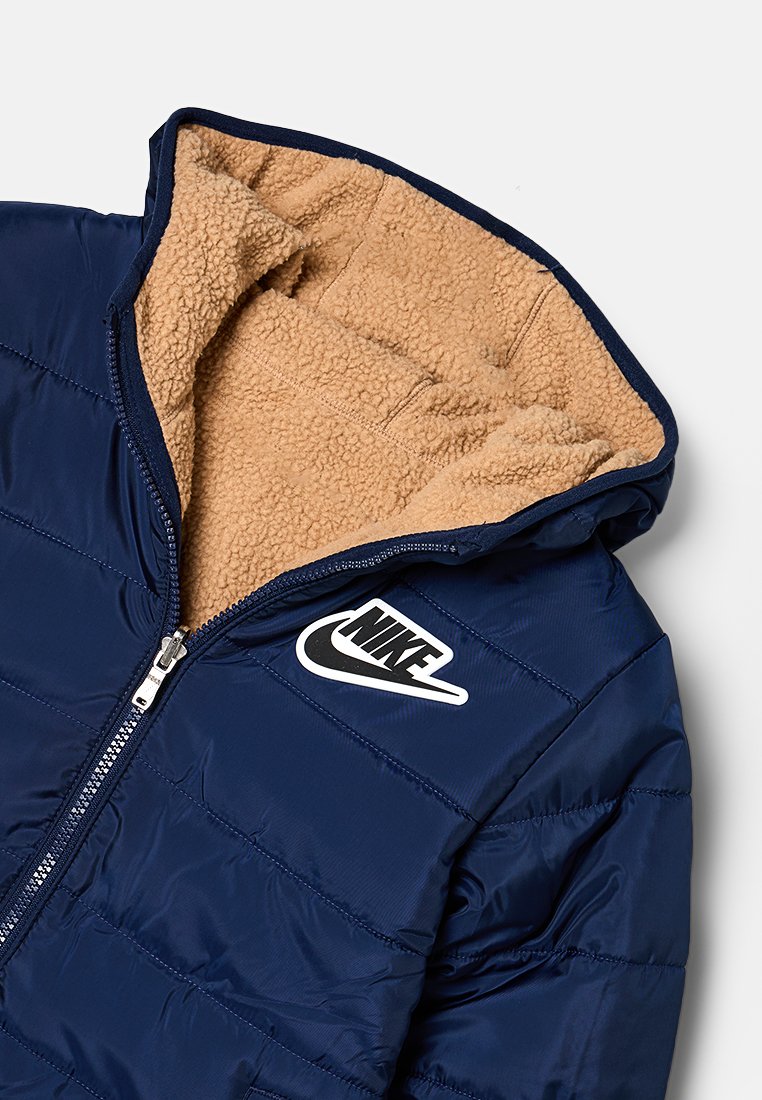 Doudoune marine avec une capuche intérieure en polaire beige. Possède une fermeture éclair à l'avant et un logo Nike blanc sur la poitrine gauche.