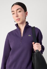 Paarse zip-up sweatshirt met een opstaande kraag en een klein groen geborduurd logo. Het model houdt een zwarte leren tote bag vast.