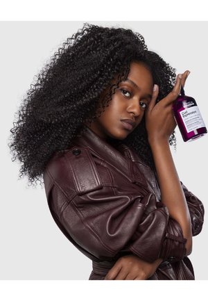 Krullend zwart haar, draagt een bordeauxleren jack. Houdt een paarse fles vast met het label "Curl Expression," dat een wit label en zwarte tekst heeft.