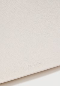 Κοντινό πλάνο υφασμένης λευκής δερμάτινης επιφάνειας με διακριτικό ανάγλυφο λογότυπο "Calvin Klein" κοντά στο κάτω άκρο.