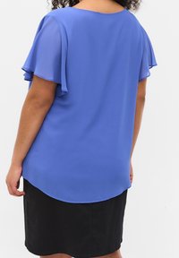 Blouse en chiffon bleu avec des manches courtes superposées et un col rond, associée à une jupe noire, dotée d'une texture lisse.