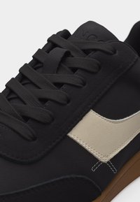 Melna sporta apavi ar melnām auklām, krēmkrāsas sānu svītru un gumijas krāsas zoli, tuvplānā, fokuss uz auklu zonu un augšējo sānu daļu.