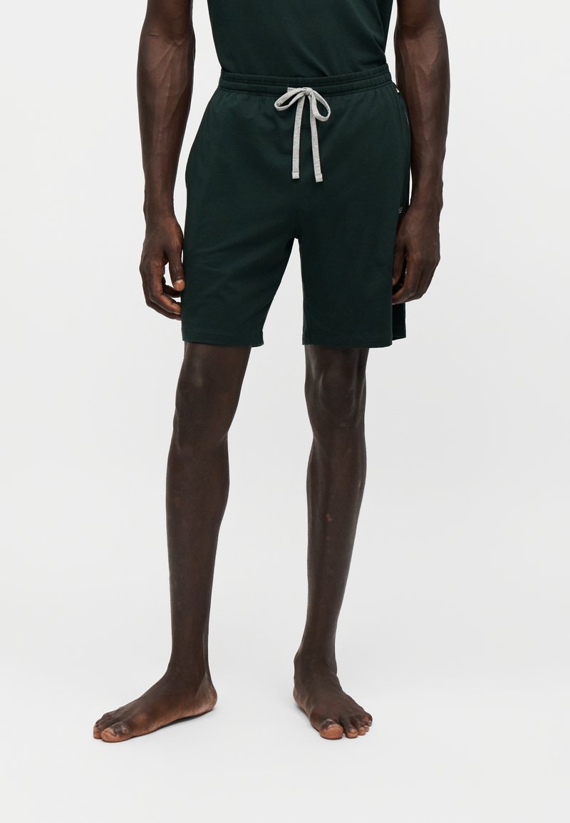 Homme portant un short noir à cordon et une chemise noire, debout pieds nus devant un fond clair uni.