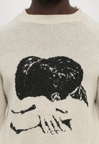 Pleteni pulover u svijetloj bež boji s crnim grafičkim dizajnom ruku i glave. Tekstura je mekana s suptilnim flekama.