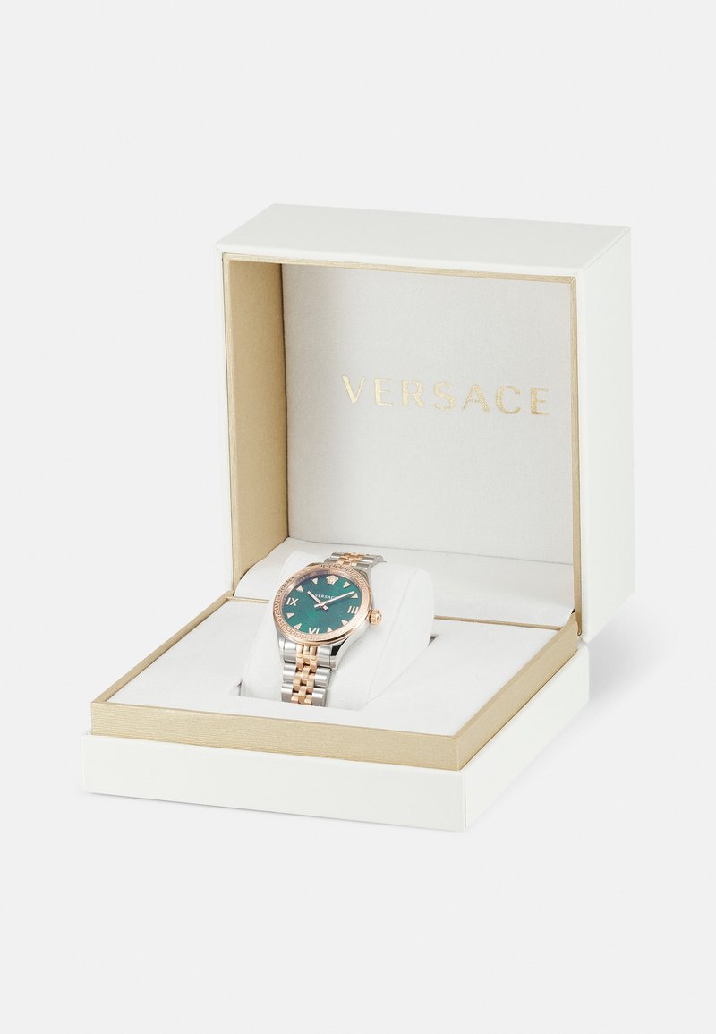 Reloj Versace en una caja blanca, con una esfera teal, acentos en oro rosa, una pulsera de plata y oro rosa, y marcadores en números romanos.