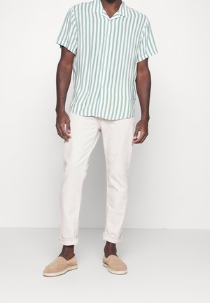 Chemise à manches courtes rayée vert et blanc, pantalon de couleur claire, et chaussures beiges faciles à enfiler, debout devant un fond uni.