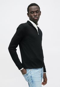 Schwarzer Reißverschluss-Pullover mit langen Ärmeln, schmaler Schnitt, weiche Textur; getragen über einem weißen Hemd mit Kragen, kombiniert mit hellblauen Jeans.