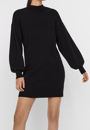 Frau in einem schwarzen, langärmligen Minikleid mit Puffärmeln und enganliegenden Bündchen, stehend vor einem schlichten hellen Hintergrund.