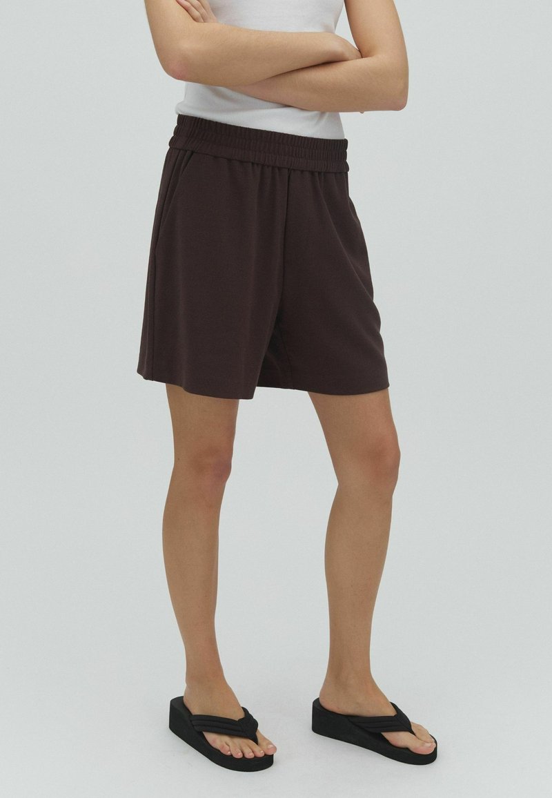 Personne portant un short marron foncé à taille élastique, un haut blanc sans manches et des tongs à plateforme noires, debout les bras croisés.
