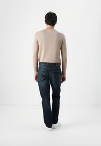 Moški obrnjeni proč v bež srajci z dolgimi rokavi, temno modrih kavbojkah in belih supergah, na enobarvni beli podlagi.