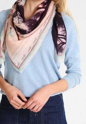 Femme portant un pull bleu clair et une jupe en denim foncé, accessoirisée d'une écharpe rose et violette à motifs autour du cou.
