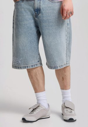 Parte inferiore del corpo di una persona che indossa pantaloncini di jeans azzurri, calzini bianchi e scarpe da ginnastica grigio chiaro con ampie linguette.
