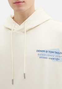 Felpa color crema con cappuccio, dotata di un cordino con punte in argento. Presenta una stampa grafica blu con il nome del marchio e l'anno di fondazione sul davanti.