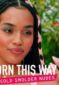 Een jonge persoon met gevlochten haar versierd met roze en zwarte linten, draagt een lichtroze top en subtiele make-up, terwijl ze glimlacht tegen een vervaagde achtergrond.