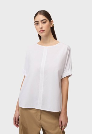 Kobieta o prostych brązowych włosach nosi białą bluzkę z krótkim rękawem z centralnym szwem, beżowe spodnie oraz złoty nausznik i pierścionki.