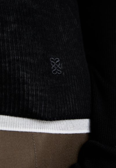 Uvećanje crne rebraste tkanine s suptilnim izvezenim logotipom iznad bijelog obruba i smeđe glatke tkanine ispod.