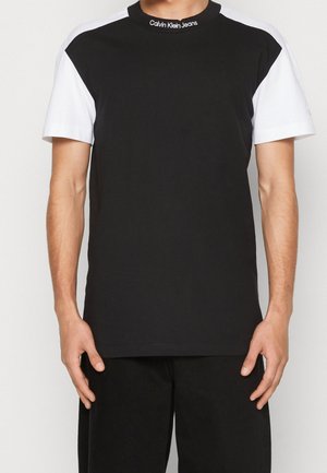 Man draagt een zwart T-shirt met witte mouwen en de tekst "Calvin Klein Jeans" op de halskraag, staand tegen een effen achtergrond.