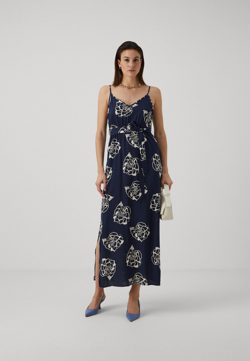 Robe maxi marine avec des motifs floraux blancs, bretelles spaghetti ajustables, taille cintrée, fente sur le côté et tissu lisse. Assortie avec des chaussures bleues.