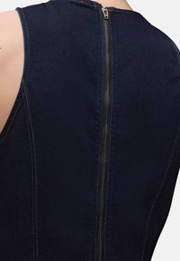 Οπίσθια όψη ενός αμάνικου σκούρου μπλε τζιν φορέματος με εκτεθειμένο μεταλλικό φερμουάρ που τρέχει κάθετα κατά μήκος του κέντρου.