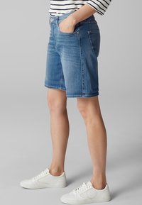Pantalones cortos de mezclilla en azul claro con un corte recto, que cuentan con bolsillos delanteros y cierre de botón. Se llevan con zapatillas blancas.