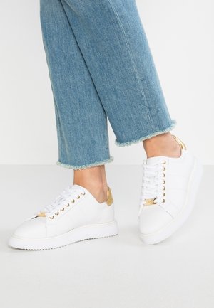 Osoba ubrana w jasnoniebieskie skrócone dżinsy i białe skórzane trampki ze złotymi akcentami, stojąca na jednolitej białej powierzchni.