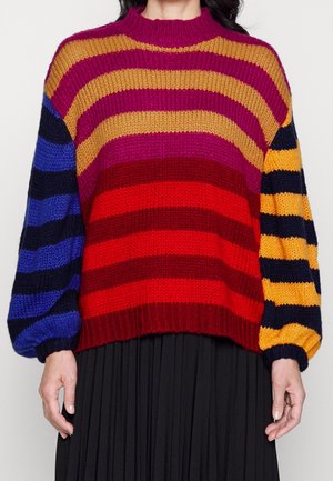 Strikket sweater med brede horisontale striber i rød, bordeaux, sennep, lilla, blå og sort, kombineret med en sort plisseret nederdel.