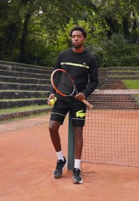 Svart hettegenser med grønn logo, svarte shorts med neongul logo, holder en tenniskølle og ball, står nær et tennisnett på leircourt.