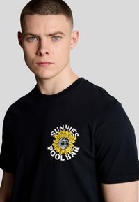 Sort bomulds T-shirt med et grafisk motiv af en smilende sol iført solbriller, med teksten "SUNNIE'S POOL BAR" i hvidt omkring den.