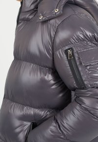 Vadderad grå jacka med en glansig yta. Har en ficka med dragkedja på ärmen och knappstängning. Det quiltade mönstret ökar isoleringen.