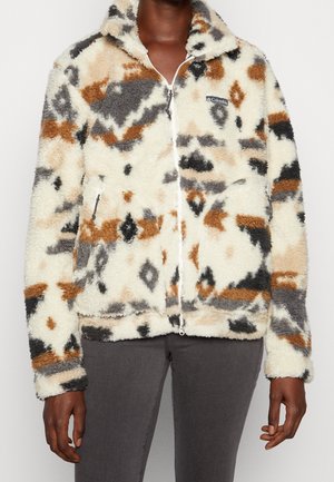 Personne portant une veste polaire zippée avec un motif abstrait marron, noir, blanc et gris et deux poches avant zippées.