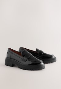 Black patent leather loafers with a low platform sole and textured tread. Straight lines and a simple strap accent the design.