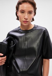 Top curto de couro preto com mangas curtas e decote redondo, textura suave e detalhes de costura visíveis. A modelo está a segurar uma clutch de couro correspondente.