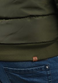 Dunkelgrüne Steppjacke mit geripptem Saum, die die strukturierte Stoffoberfläche und ein Lederlogo-Patch zeigt. Getragen zu blauen Denim-Jeans.