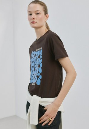 Junge Frau mit blonden Haaren im Pferdeschwanz trägt ein schwarzes T-Shirt mit blauem Blumenmuster und Schriftzug, kombiniert mit einem weißen Pullover, der um die Taille gebunden ist.