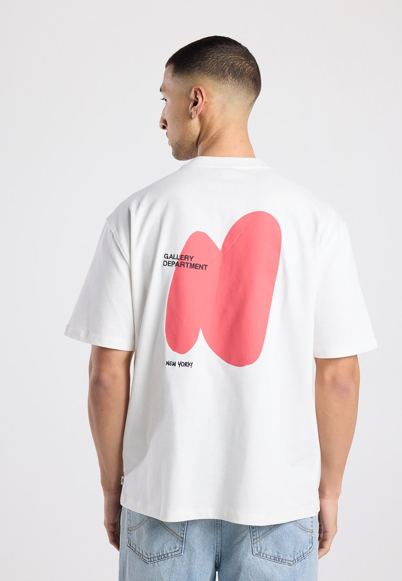 Man draagt wit T-shirt met grote abstracte roze vorm en tekst "GALLERY DEPARTMENT" en "NEW YORK!" aan de achterkant, gecombineerd met lichtblauwe spijkerbroeken.