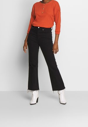 Oranje geribd shirt met lange mouwen en knoopdetails, gecombineerd met zwarte flared jeans met rafelige zomen en witte enkellaarsjes.