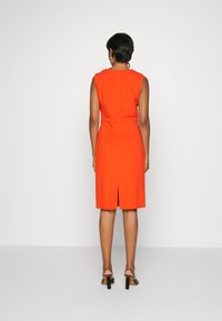 Ärmlös orange klänning med en figurnära design, bakre dragkedjestängning och mittslitsdetalj vid fållen, kombinerad med svarta klacksandaler med blockliknande klack.