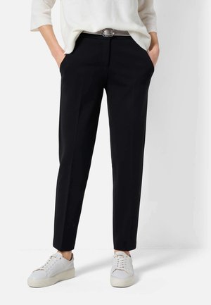 Personne portant un pantalon noir sur mesure, des baskets blanches et un pull blanc, les mains dans les poches, debout devant un fond uni.