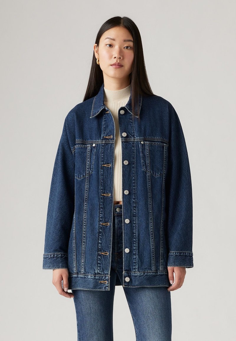 Jachetă din denim de culoare albastru închis, cu închidere cu nasturi în față, două buzunare pe piept și o croială lejeră. Material texturat cu detalii de cusătură.