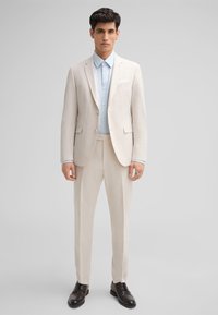 Hellebeiges Anzug mit taillierter Passform, strukturiertem Stoff, einreihigem Blazer, kombiniert mit einem hellblauen Hemd und schwarzen Schuhen.