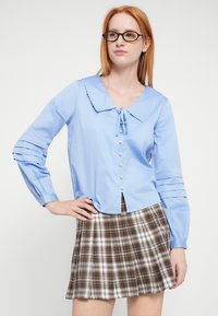 Jasnoniebieska bluzka z falbankowanym kołnierzem, wiązana z przodu, z perłowymi guzikami. Posiada warstwowe, marszczone rękawy. Połączona z brązową spódnicą w kratę.